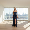 A woman modeling a strapless navy blue maxi dress with a sweetheart neckline, a ruffled waist detail, and a high thigh slit, set against a plain white background.