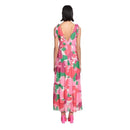 A woman viewed from the back wearing a sleeveless maxi dress with a pink, green, and white abstract floral pattern and a deep V-back, paired with bright pink high heels on a white background.