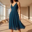 A woman modeling a sleeveless teal blue midi dress with a V-neckline and a pleated tulle skirt, paired with black strappy high-heeled sandals against a plain white background.