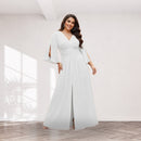 A woman models a floor-length white dress with a wrap-style bodice, flared sleeves, and a front slit in a bright indoor room with pale walls and wood flooring.