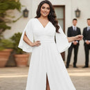 A woman wearing a white floor-length gown with wide flared sleeves and a V-neckline, standing outdoors in front of a building with two blurred men in suits behind her.
