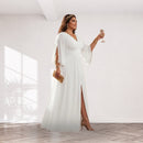 A woman models a floor-length white gown with wide draped sleeves, a V-neckline, and a thigh-high slit, standing in a bright minimalist room and holding a champagne flute.