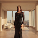 A woman modeling a floor-length black sequined gown with long sleeves, a boat neckline, and a high front slit, photographed against a plain white background.