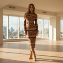 A woman modeling a long-sleeve midi dress with a horizontal desert landscape print in shades of terracotta, brown, beige, and green, paired with tan cowboy boots, against a plain white background.