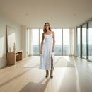 A woman modeling a sleeveless white maxi dress with a light blue abstract print and a front slit, paired with tan cowboy boots, against a plain white background.