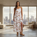 A woman modeling a sleeveless white maxi dress with a cowboy and cattle print, featuring thin straps, a high side slit, and brown cowboy boots against a plain white background.